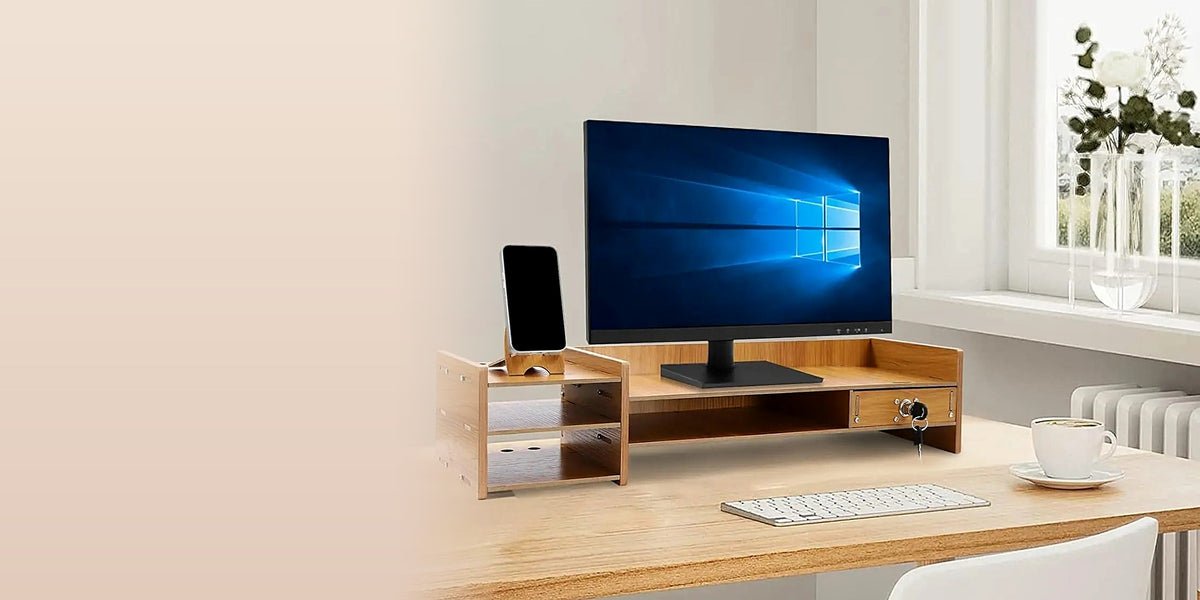 Solid wood monitor riser with a phone dock and locking drawer on a light oak desk beside a sunlit window